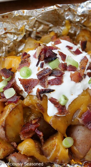 A close up of an open potato foli packet recipe with potatoes, bacon, sour cream, green onions.