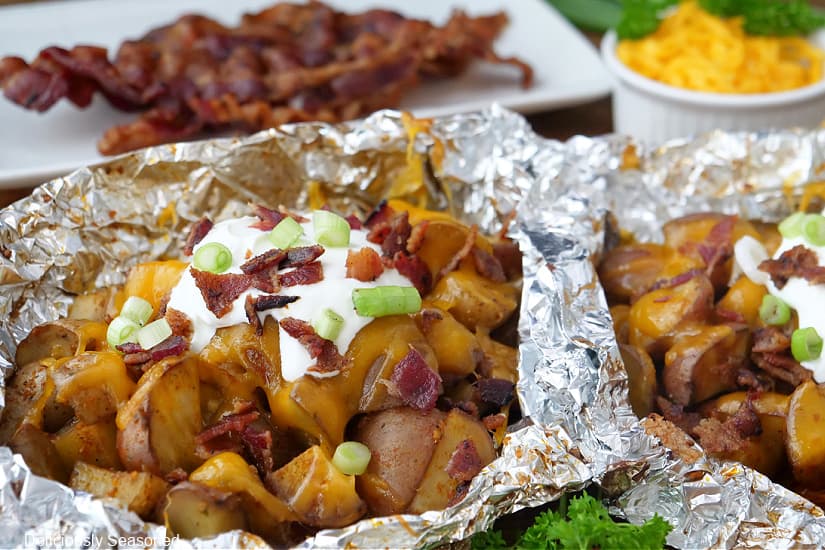 A horizontal photo of potatoes in foil packets topped with cheese, sour cream, bacon and green onions.