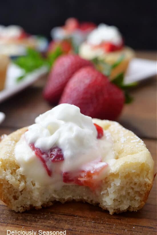 A pancake cup of a wood surface with a bite taken out of it showing the cream cheese filling and fresh diced strawberries and whipped cream on top.