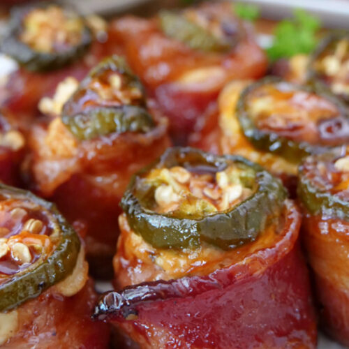 A close up of a plate full of smoked pig shots which are bacon wrapped around sausage, filled with cheese mixture and a jalapeno slice on top.