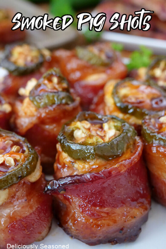 A close up of a plate full of smoked pig shots which are bacon wrapped around sausage, filled with cheese mixture and a jalapeno slice on top.