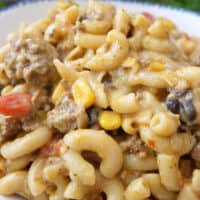 A close up of a white bowl filled with a serving of ground beef, pasta, beans and corm, and more in a creamy sauce.