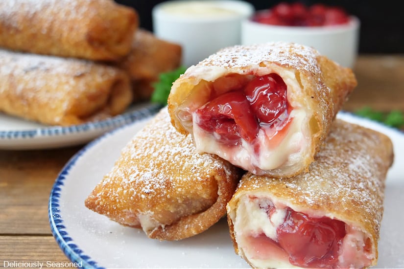 Two whtie plates with blue trim with three egg rolls each on them with two eggs with bites out of them showing the cherry cream cheese filling.