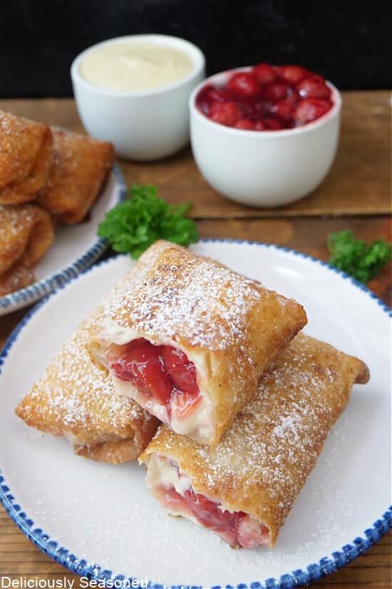 Cherry egg rolls on a white plate with blue trim with two small white bowls in the background filled with cherries and one with cream cheese.