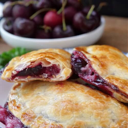 A white plate with blue trim with three cherry empanadas on it with a white bowl filled with fresh cherries.