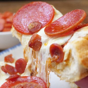 A slice of homemade pizza bread lifted off a white plate showing the stretchy cheese and the different types of pepperonis.