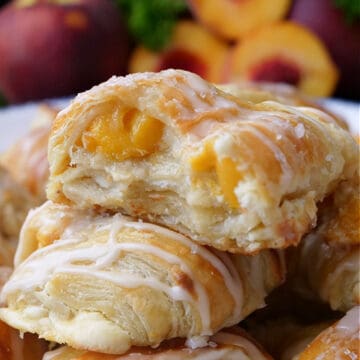 A stack of peach puff pastries on a white plate with fresh peaches in the background.