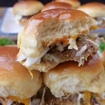 Three pulled pork sliders on a white plate with more sliders in the background.