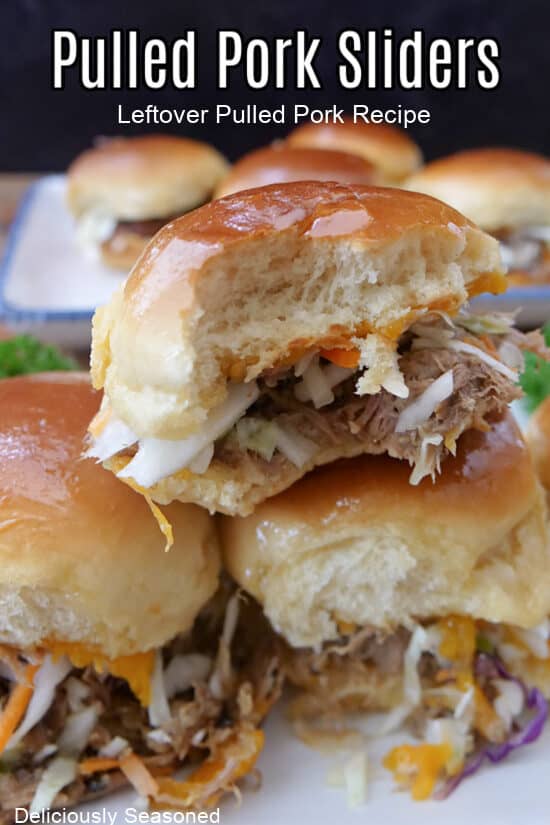 Three pulled pork sliders on a white plate with more sliders in the background.