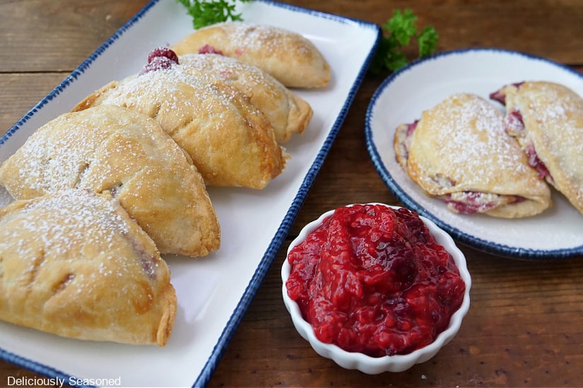 Two white plates with blue trim with raspberry empanadas on it with a small bowl of raspberry filling.