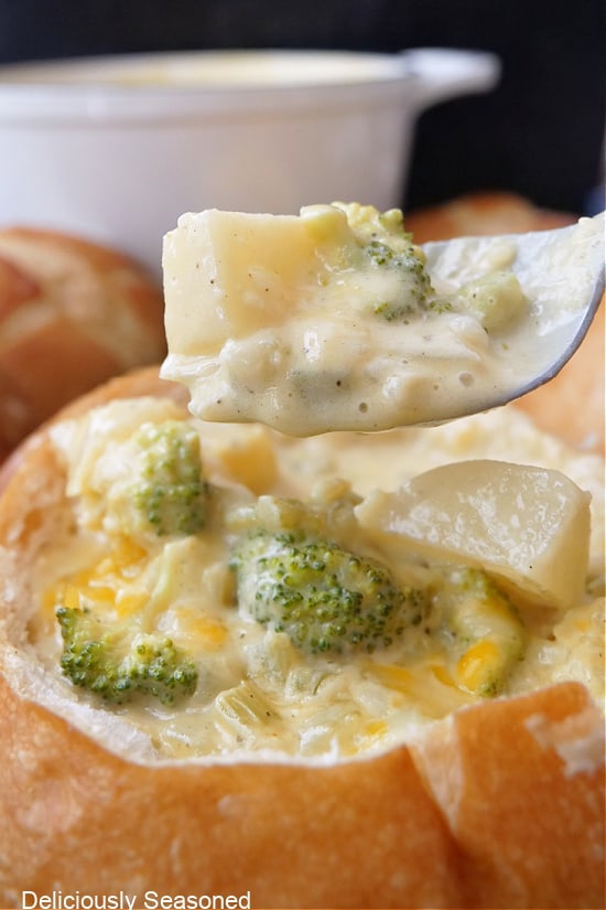 A spoon filled with potato broccoli soup on it held about a bread bowl filled with soup.