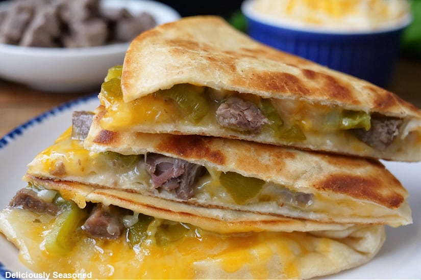 A horizontal photo of a white plate with blue trim with three pieces of a quesadilla filled with hatch chiles, steak and cheese.