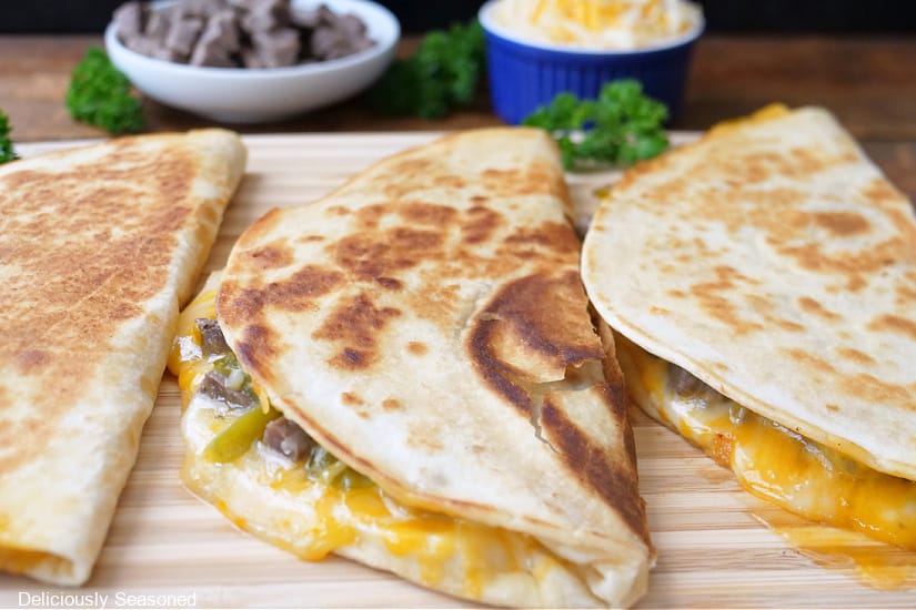 Three half quesadillas on a wood cutting board showing the melty cheese, meat, and chiles before being cut into pieces.