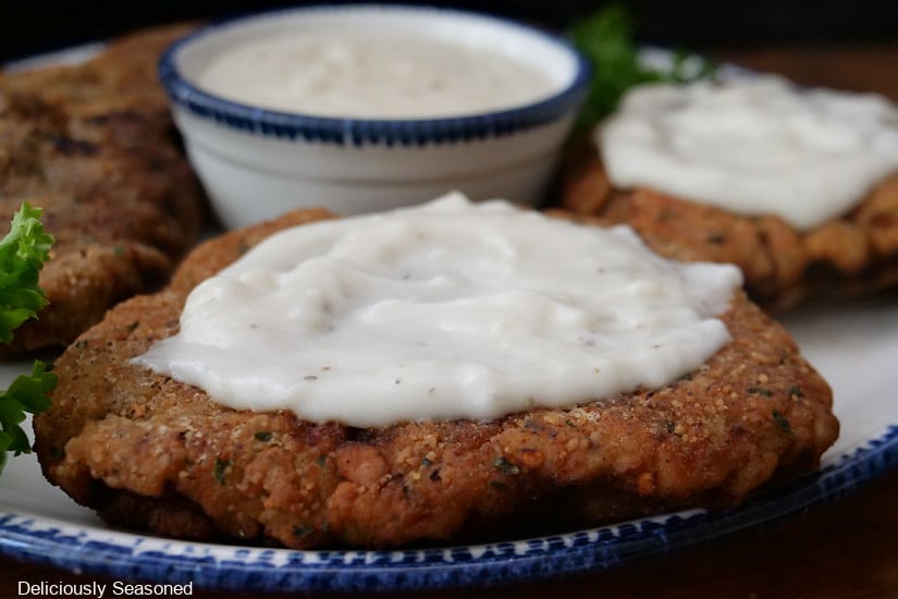 A white plate with blue trim with three chicken fried steaks on it and a bowl of white gravy.