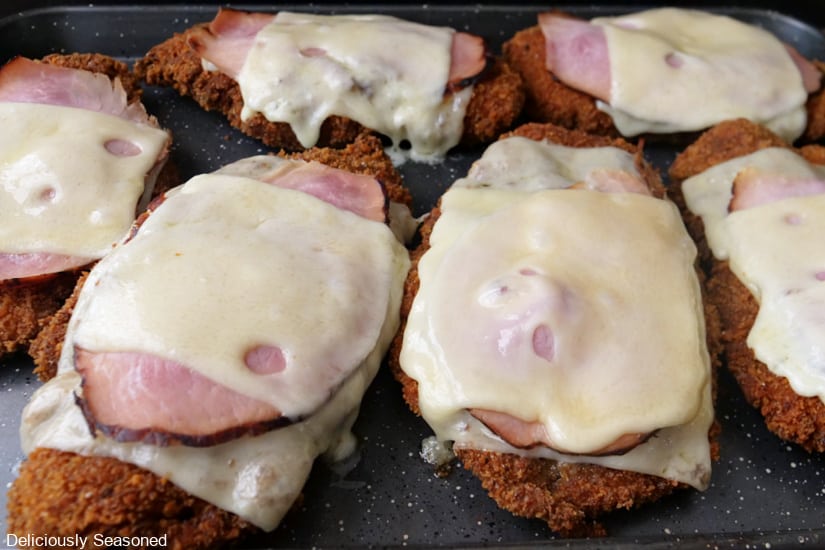 A horizontal photo of a baking sheet with six pieces of boneless fried chicken breasts with sliced ham and Swiss cheese on it.
