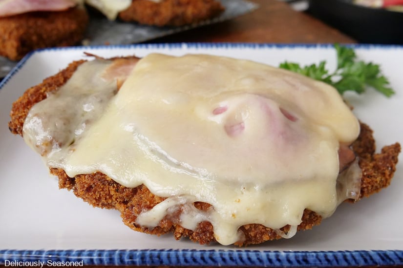 A horizontal photo of a white rectangle plate with blue trim with a piece of fried boneless chicken breasts with melted Swiss cheese and ham on it.