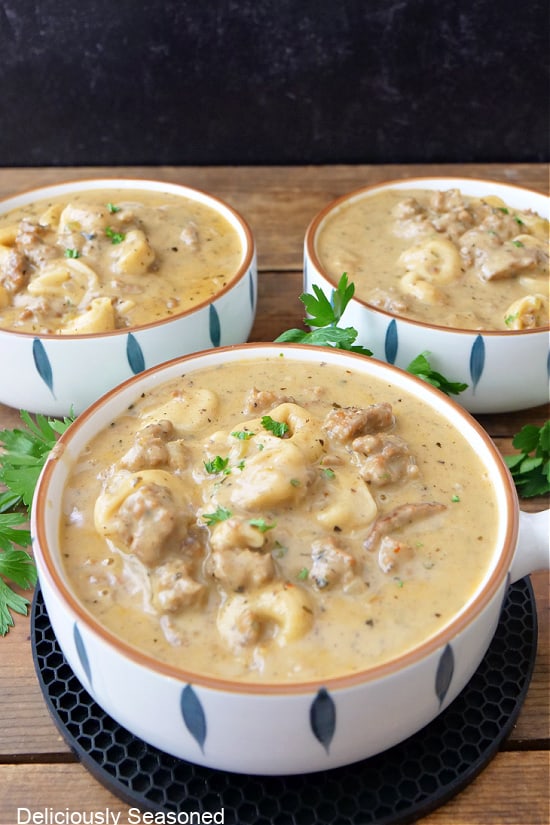 A wood surface with three white soup bowls filled with creamy tortellini soup with Italian sausage.