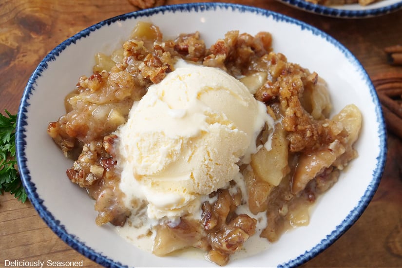 A horizontal photo of apple crumble with vanilla ice cream on top in a white bowl with blue trim.