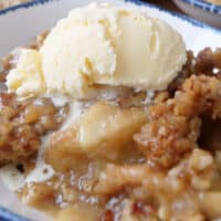 A close up of apple crumble, in a white bowl with blue trim, with a scoop of vanilla ice cream on top.