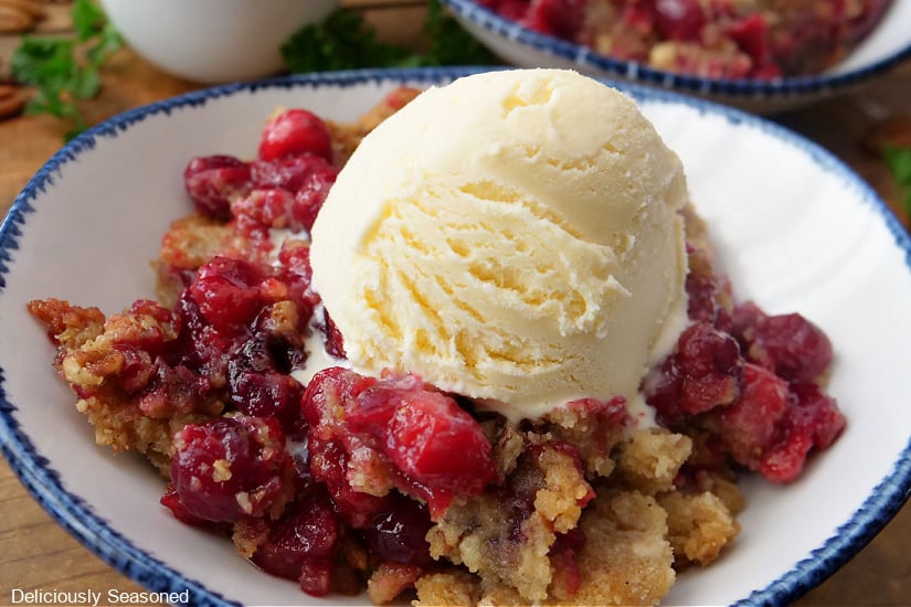 A horizontal photo of a white bowl with blue trim with a scoop of vanilla ice cream on top of cranberry crumble.