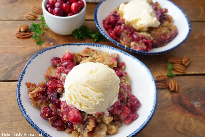 A wooden surface with two white bowls with blue trim filled with cranberry crumble topped with vanilla ice cream.