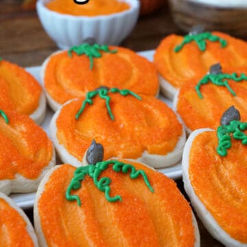 A wood surface and white plate with pumpkin shaped sugar cookies frosted with orange frosting.