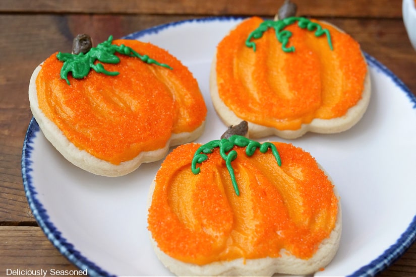 Three pumpkin shaped sugar cookies with orange frosting on a white plate with blue trim.