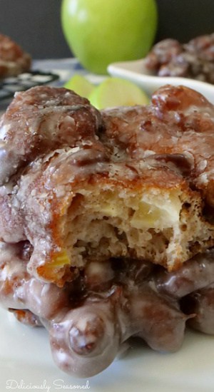 A close up shot of two apple fritters with a bite taken out of one of them, both placed on a white plate with a green apple in the background and more apple fritters as well.