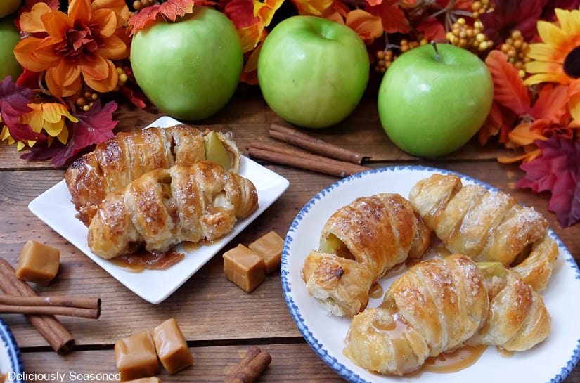 A horizontal photo of two white plates with caramel apple slices on them with green apples and fall foliage in the background.