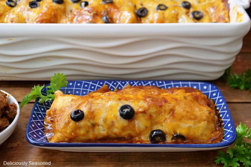 A single enchilada on a blue plate with the large white baking dish filled with more enchiladas.