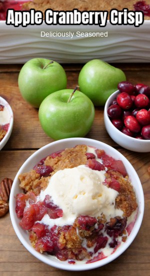 An overhead photo of a wood surface with a couple white bowls, one filled with a serving of apple cranberry crisp with vanilla ice cream and one filled with fresh cranberries, and 3 green apples are on the wood surface.
