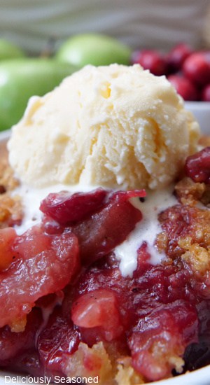 A close up of apple cranberry crisp in a white bowl with a scoop of vanilla ice cream on top.