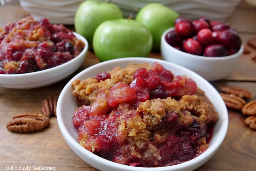 Two white bowls with servings of apple cranberry crisp in them and a small with bowl filled with fresh cranberries. Pecans and green apples in the background.