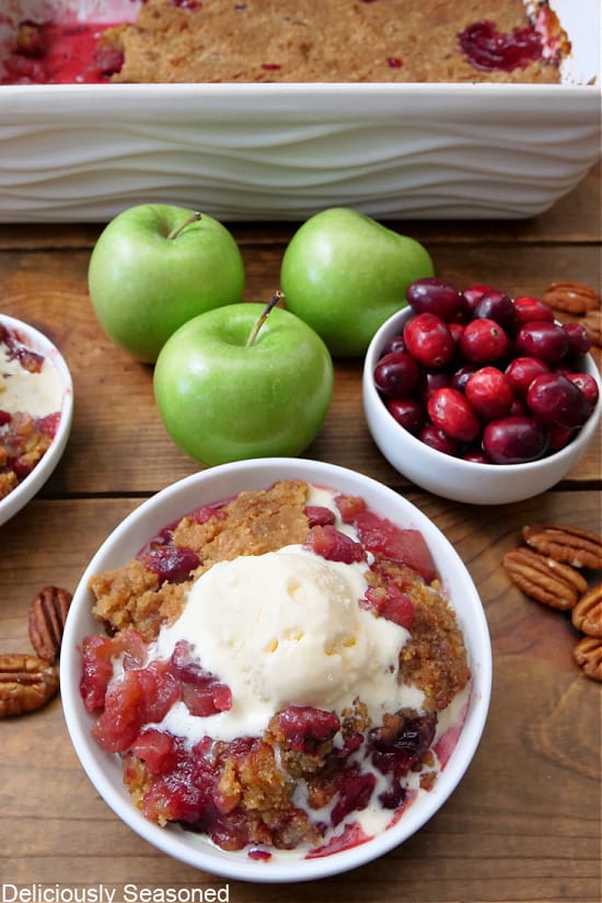 A photo of a wood surface with a white bowls, one filled with a serving of apple cranberry crisp with vanilla ice cream and one filled with fresh cranberries, and 3 green apples are on the wood surface.