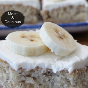 A close up photo of a banana bar on a white plate with cream cheese frosting and two slices of bananas on top of the frosting.