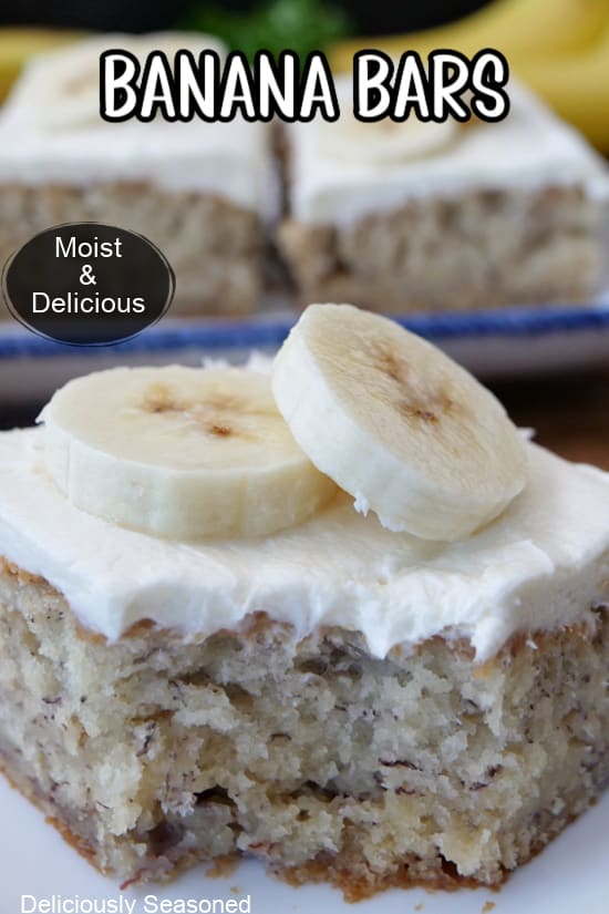 A close up photo of a banana bar on a white plate with cream cheese frosting and two slices of bananas on top of the frosting.