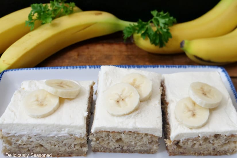 A horizontal photo of three pieces of banana bars on a white rectangle plate with blue trim with four whole bananas in the background.