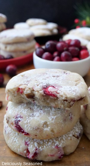 A stack of three cranberry pecan cookies on a wood surface with more cookies in the background and a white bowl filled with fresh cranberries.