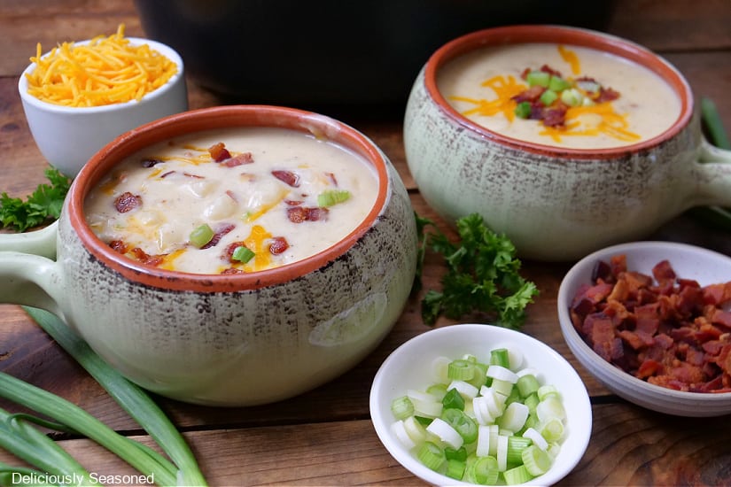 A horizontal photo of a wood surface with two soup bowls filled with potato soup, and three small bowls with cheese, green onions, and crispy bacon in them.