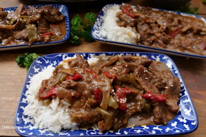 A horizontal photo of slow cooked flank steak and peppers with white rice on blue and white small rectangle plates.