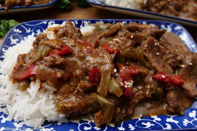 A horizontal photo of a blue and white rectangle plate filled with a serving of steak and peppers with white rice.
