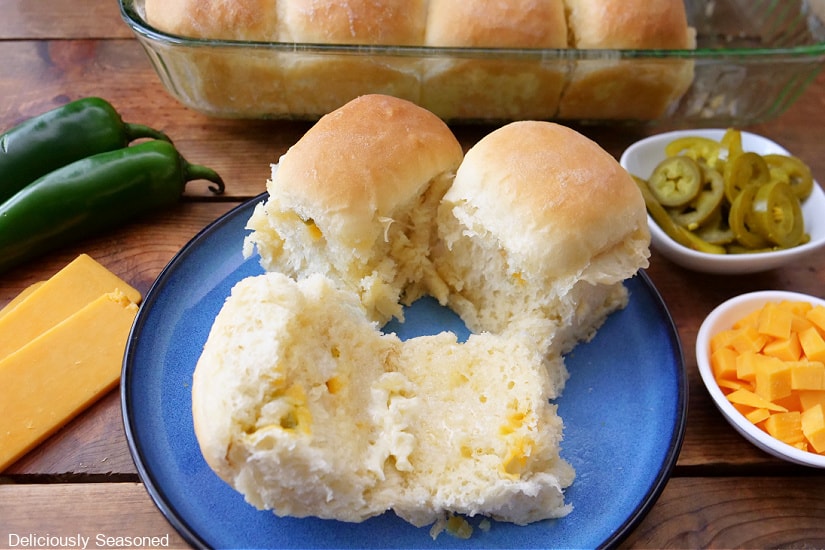 A horizontal photo of a blue plate with three dinner rolls on it with one cut in half with butter added, and cheddar cheese and jalapenos placed on a wood surface next to the plate.