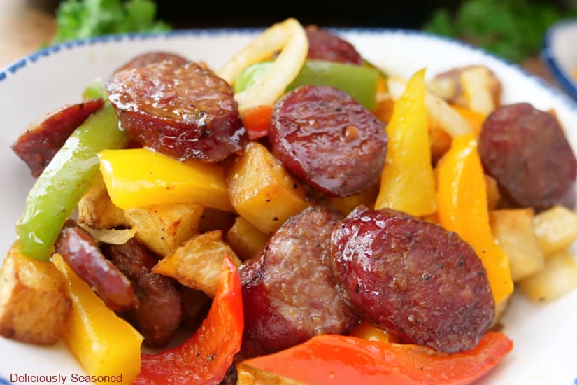 A horizontal photo of a white bowl filled with slices of smoked sausage, bell peppers, onions, and potatoes.
