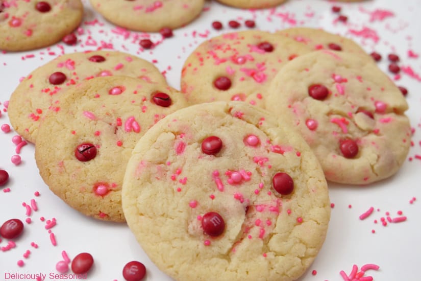 A whit surface with with sugar cookies on it with candy sprinkles and mini red M&Ms scattered around the cookies on the white surface.