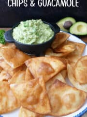 A large white plate with blue trim with homemade deep fried tortilla chips on it with a black bowl filled with homemade guacamole.