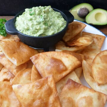 A large white plate with blue trim with homemade deep fried tortilla chips on it with a black bowl filled with homemade guacamole.