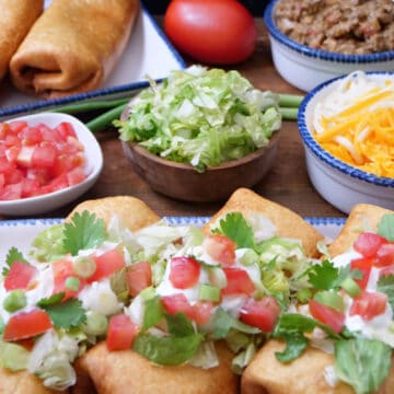A wood surface with deep fried burritos on white plates with blue trim, with lettuce, tomatoes, shredded cheese, and ground beef mixture in the background.