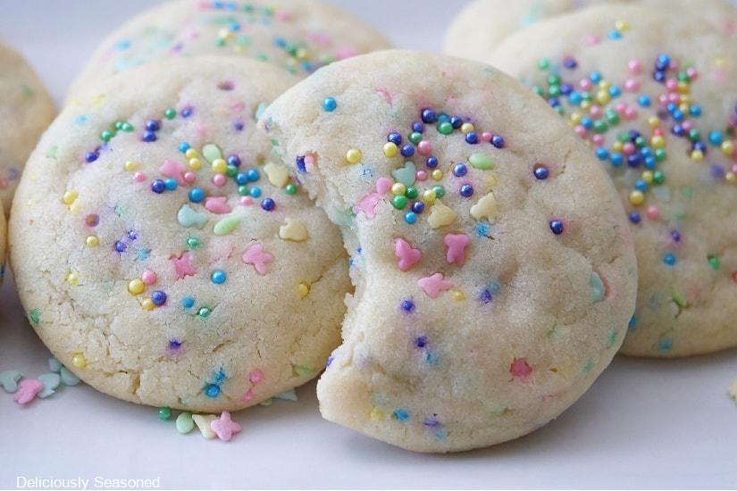 A few sugar cookies with Easter sprinkles are on a white plate and one of the cookies have a bite taken out of it.