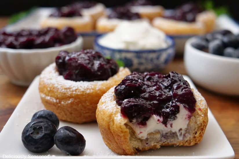 Two mini blueberry crescent cups of a white plate with a bite taken out of one of them, and more mini cups in the background.