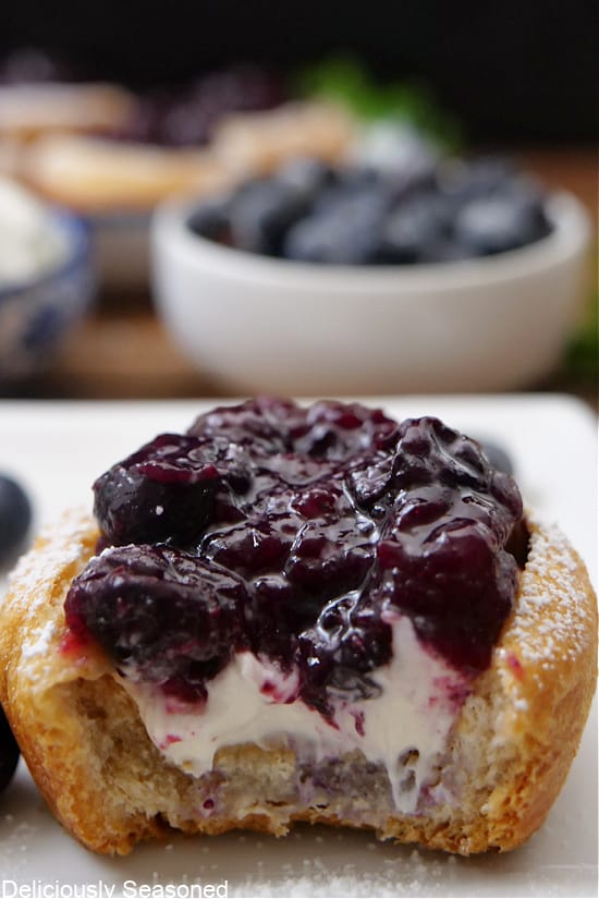 A close up of a mini blueberry crescent cup filled with cream cheese mixture and topped with blueberries with a bite taken out of it.